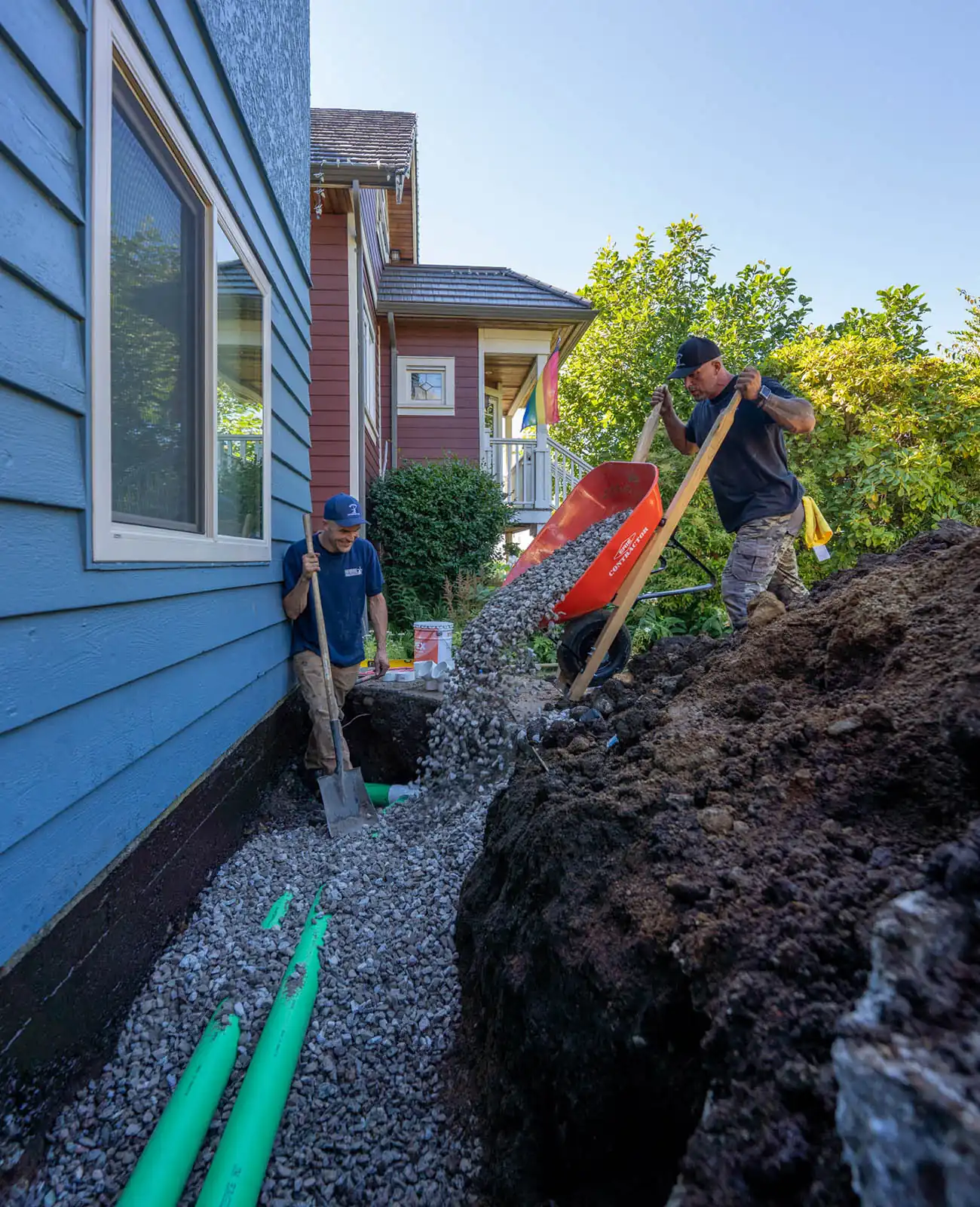 Two Modern Drainage crew members backfilling an open perimeter trench with drain rock along a blue-sided house foundation.