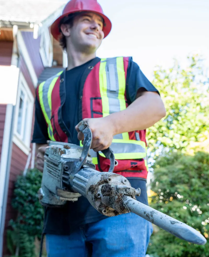 A Modern Drainage crew member holding an electric breaker with a muddy, root-fouled pipe fitting still attached to the bit, beside a house foundation.
