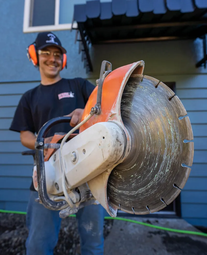 A Modern Drainage crew member operating a gas cut-off saw into a concrete slab beside a house, with a hose spraying water over the blade.