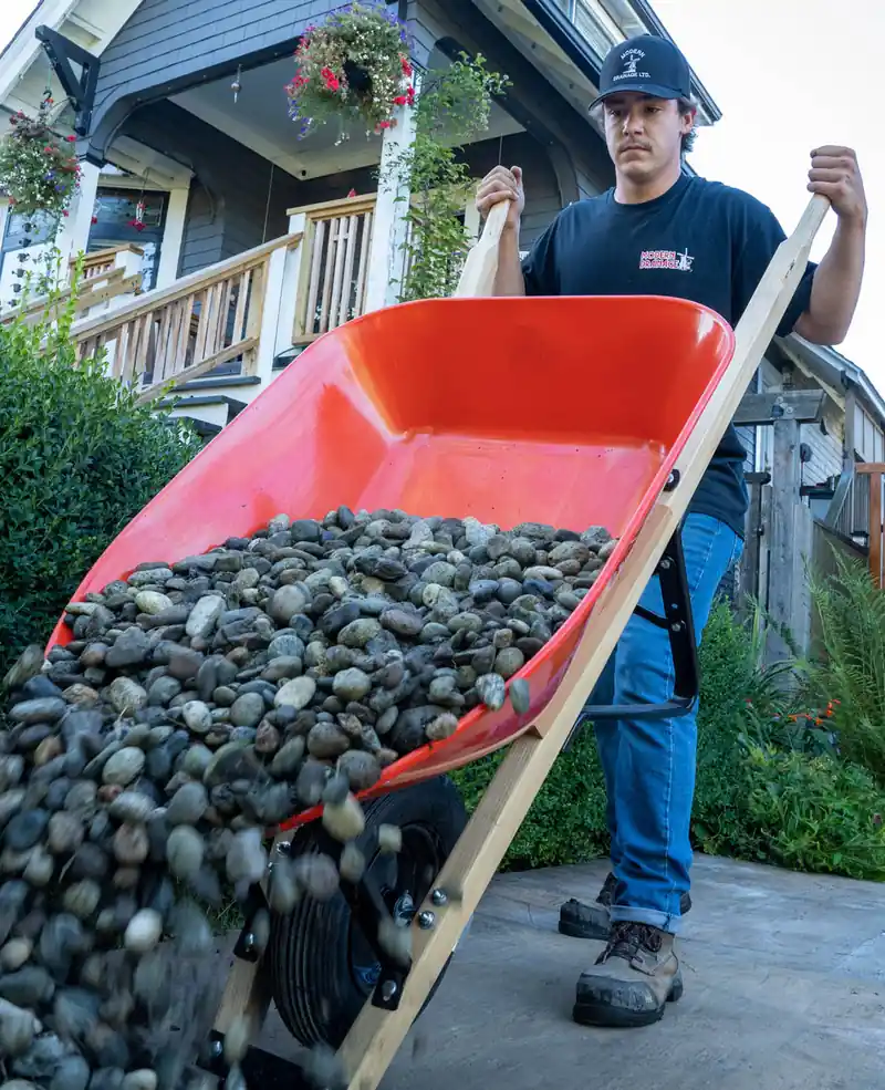 A Modern Drainage crew member tipping a wheelbarrow of drain rock down a driveway toward a house.