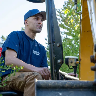 A Modern Drainage crew member seated at the controls of a compact excavator on a residential job site.