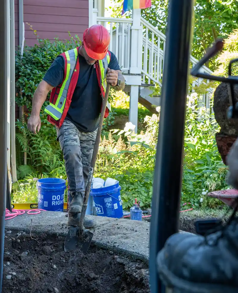 A Modern Drainage crew member hand-digging an excavation beside a house foundation, blue buckets labelled 'Do It Right' nearby.