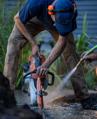 Two Modern Drainage crew members working together at dusk — one operating a cut-off saw into concrete, the other spraying water over the cut from a hose.