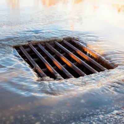 A square storm catch basin grate, submerged under standing water flowing across pavement after rain.