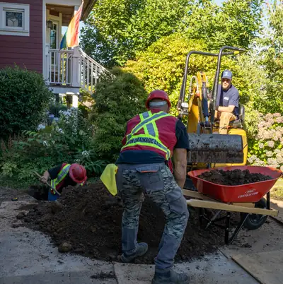 Two Modern Drainage crew members and a compact excavator working a large open trench beside a house's front yard.