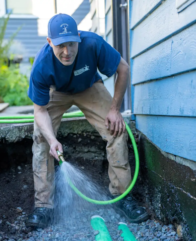 A Modern Drainage crew member flushing a newly laid section of green drain pipe with a hose, in an open trench along a blue-sided house foundation.