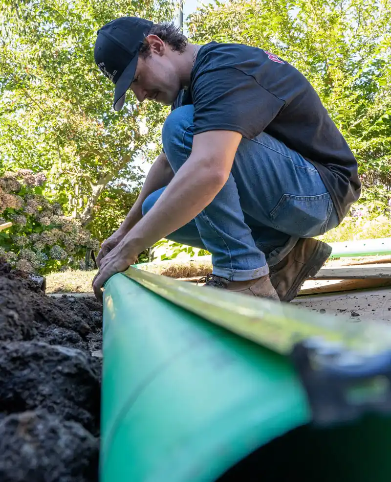 An open trench along a North Vancouver home's foundation, green perimeter drain pipe being set into gravel bedding before the ground closes back over it.