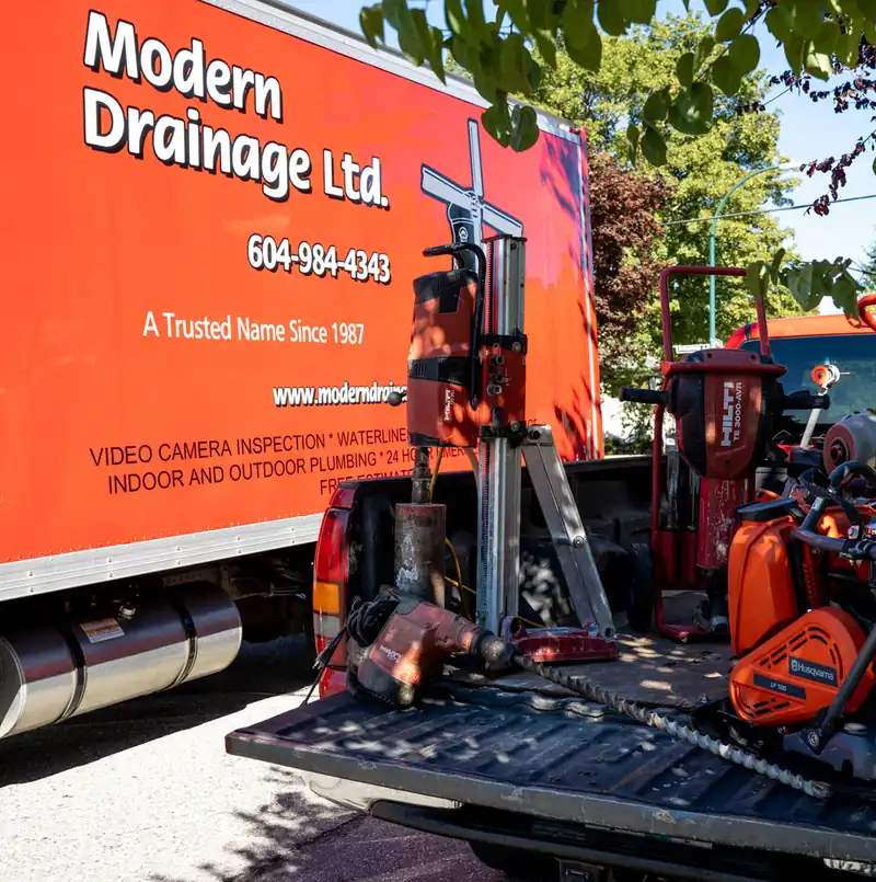 The side of a Modern Drainage service truck reading 'Video Camera Inspection, Waterline, Indoor and Outdoor Plumbing, 24 Hour Emer...' beside a truck bed loaded with a core drill and breaker tools.