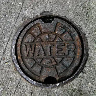A close-up of a cast-iron 'WATER' service cover set into a concrete sidewalk.